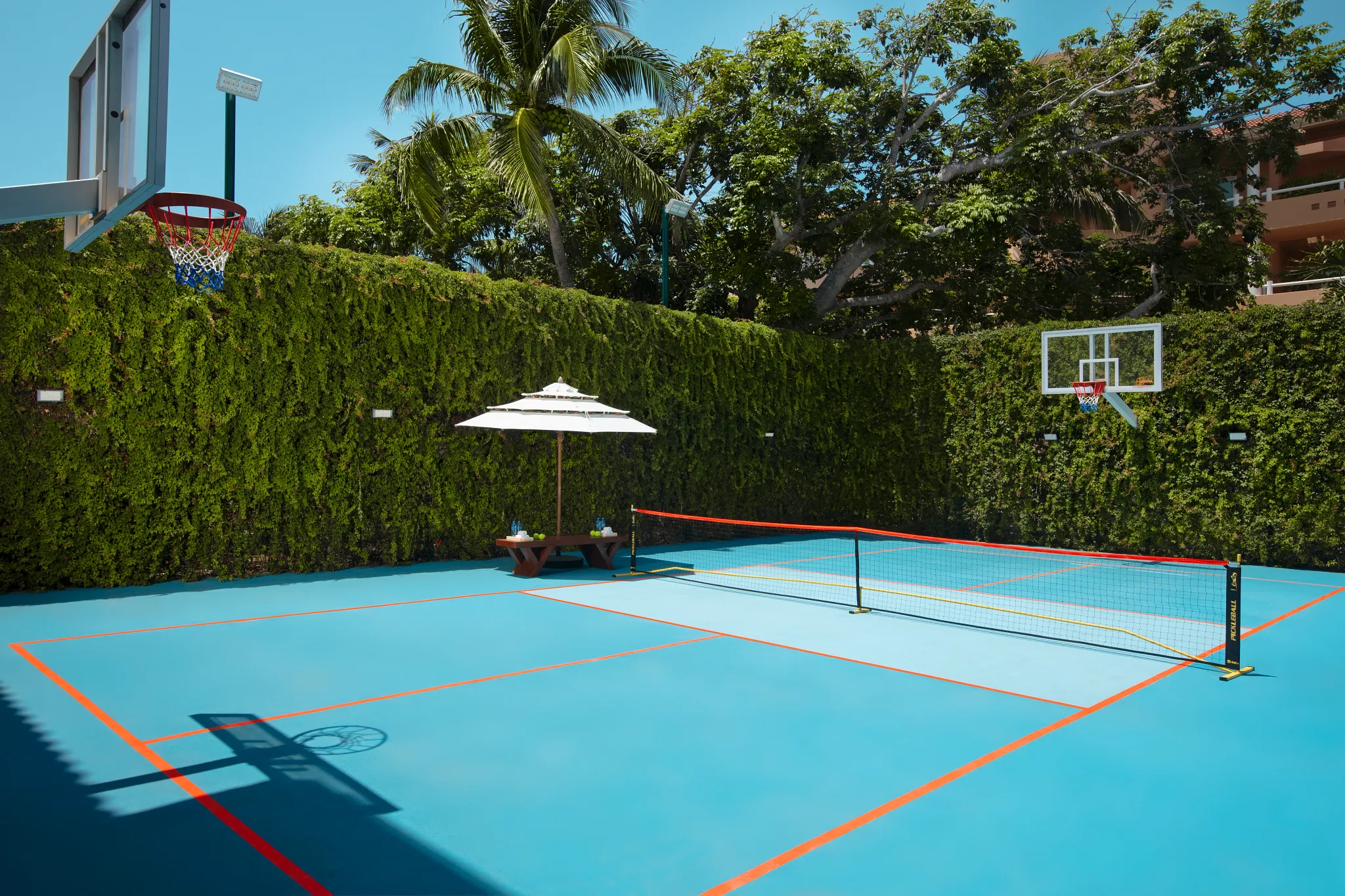 Outdoor pickleball court with basketball hoops at Dreams Aventuras Riviera Maya, surrounded by lush greenery and tropical landscaping.