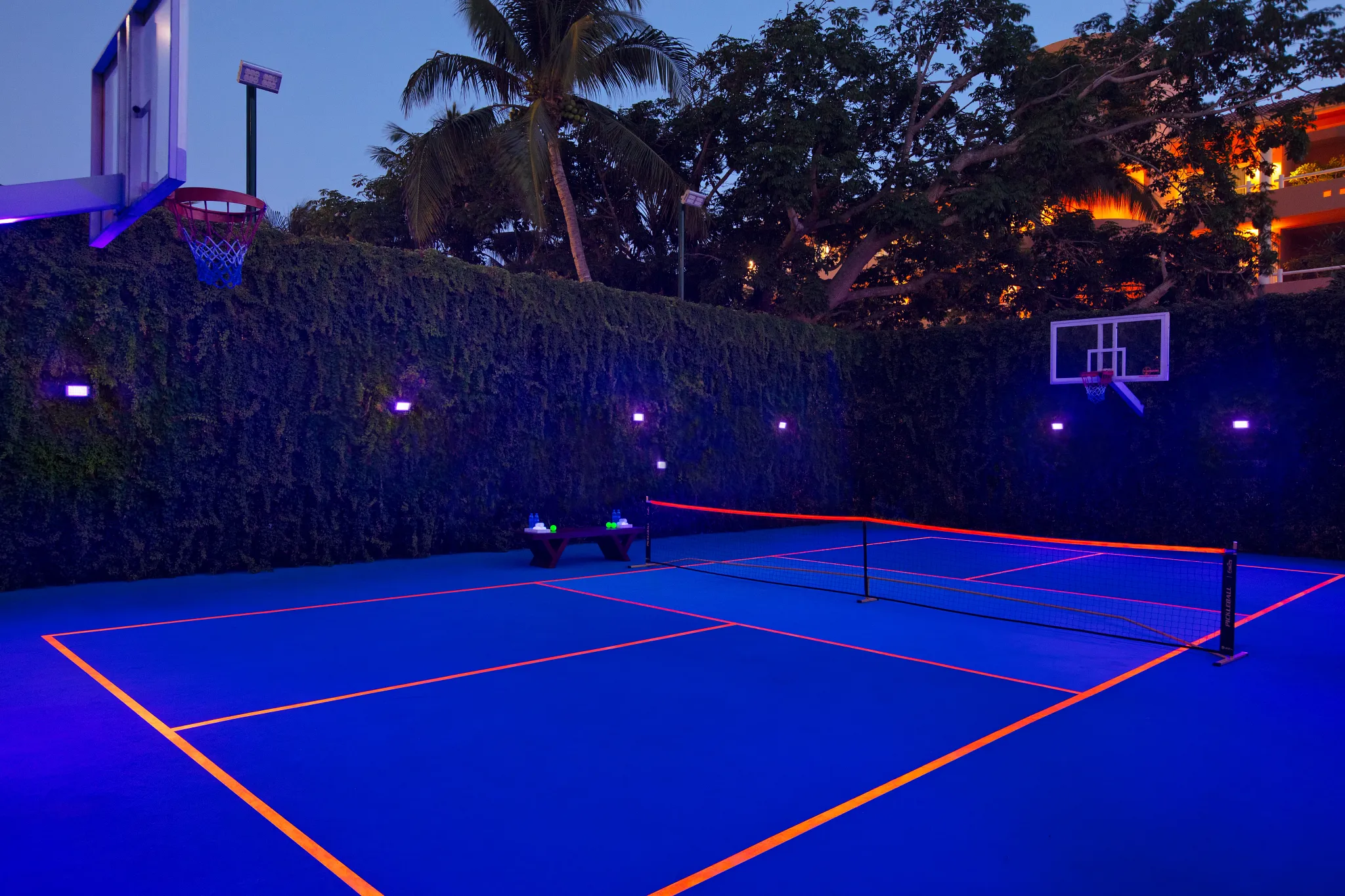 Outdoor pickleball court illuminated at night with neon lines and basketball hoops at Dreams Aventuras Riviera Maya.